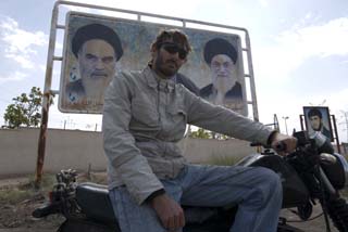 Matthew VanDyke on his MZ Kanuni motorcycle in front of a Ayatollah Khomeini and Khamenei billboard in Iran