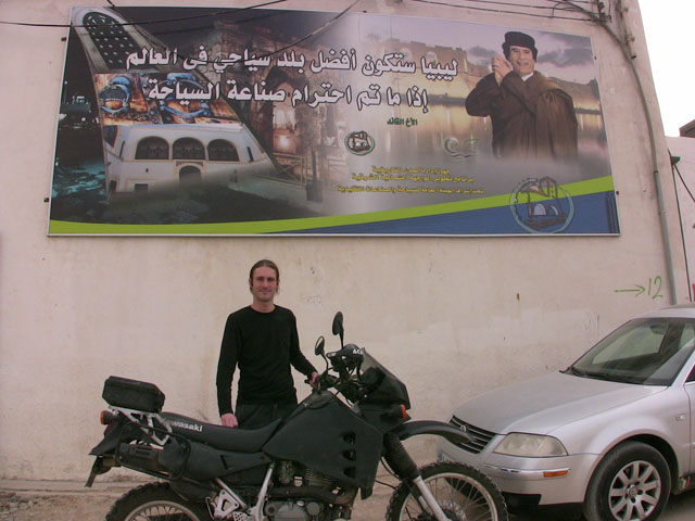 Matthew VanDyke with his Kawasaki KLR650 motorcycle in front of a poster of Muammar Gaddafi in Tripoli Libya