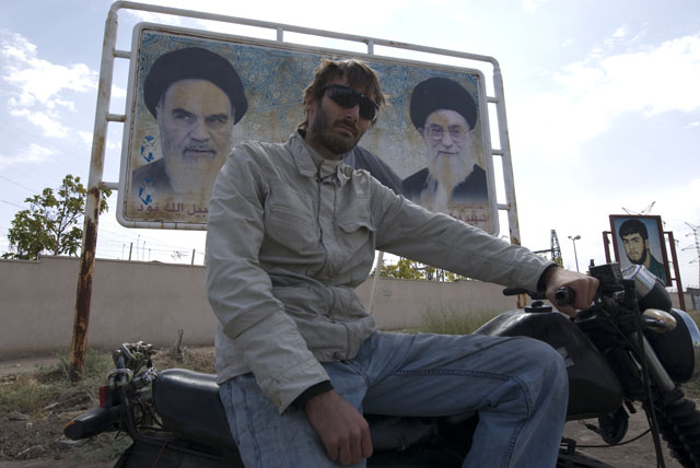 Matthew VanDyke on his MZ Kanuni motorcycle next to a billboard of Ayatollahs Khomeini and Khamenei in Iran